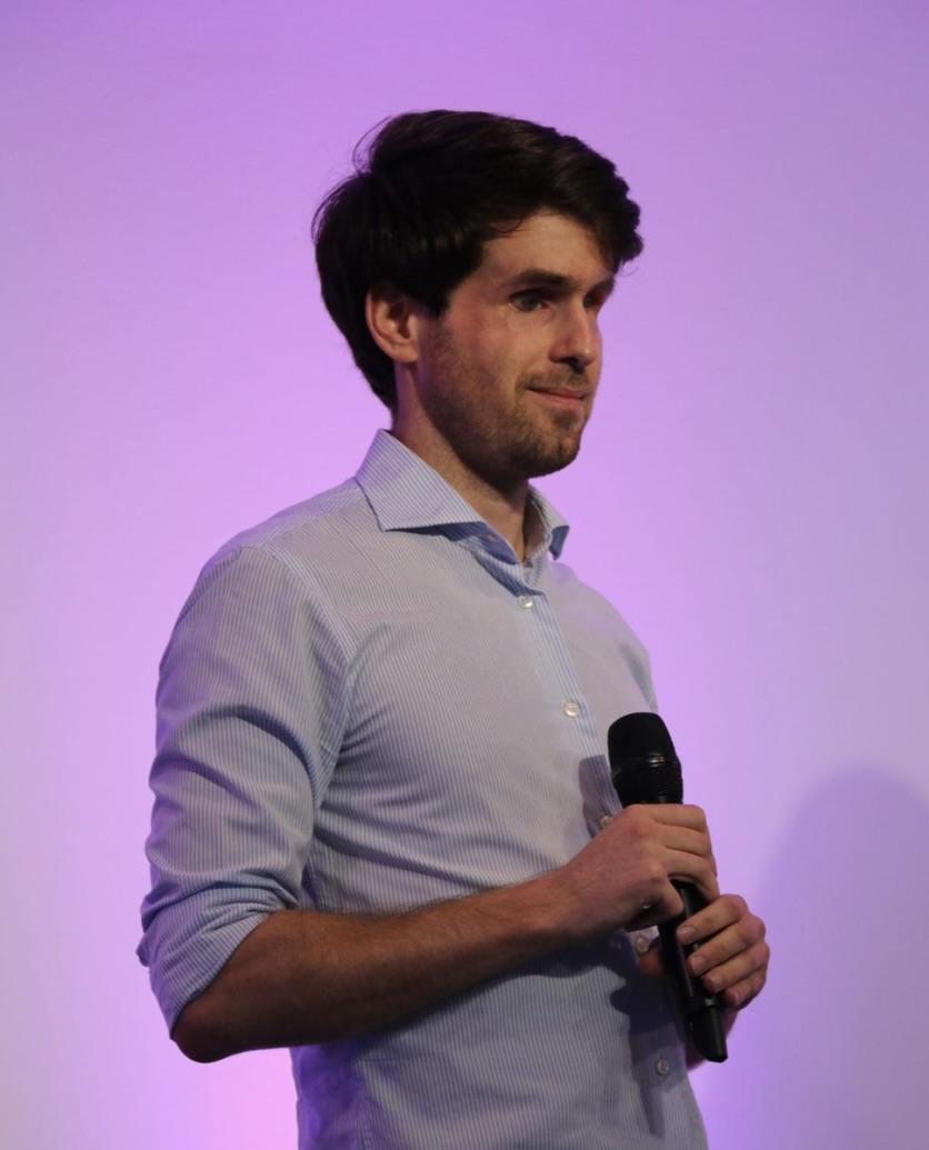 Portrait de Thibault Trancard, jeune homme les cheveux brun portant une chemise bleu qui tient un micro