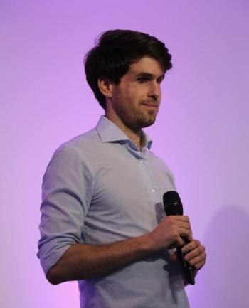 Portrait de Thibault Trancard, jeune homme les cheveux brun portant une chemise bleu qui tient un micro